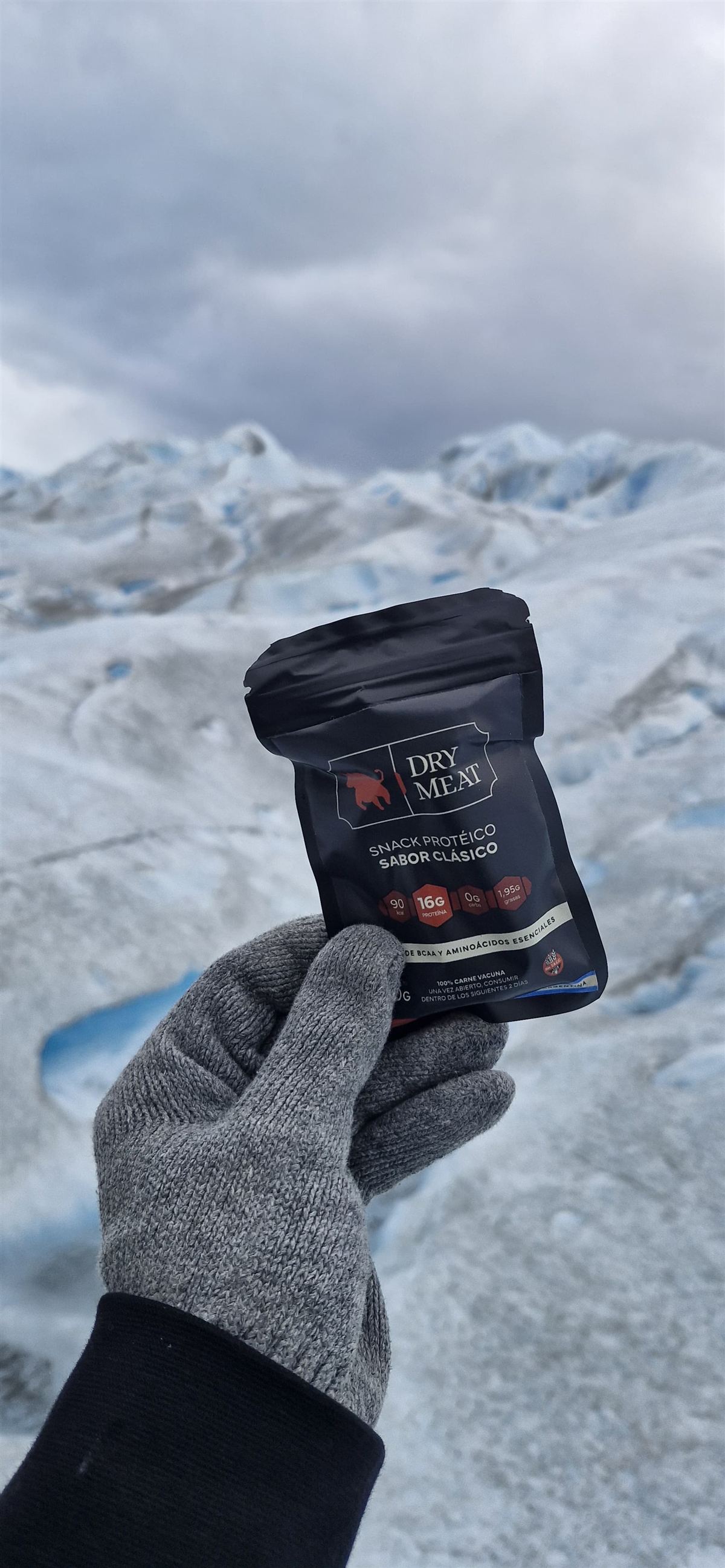 Una mano con guante sosteniendo un pack de DryMeat frente a un glaciar